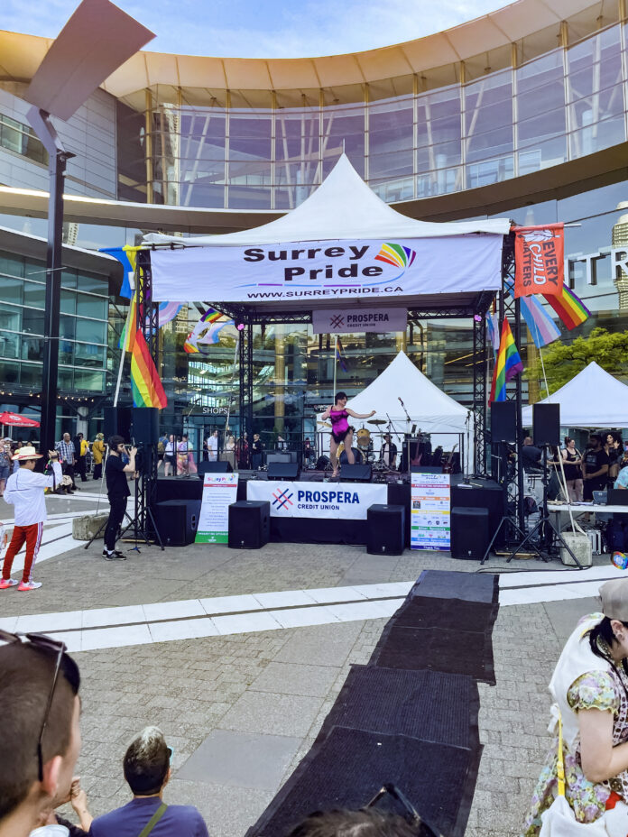The photo is of the outdoor Surrey Pride Festival. A drag performer is on an outdoor stage as the crowd watches.