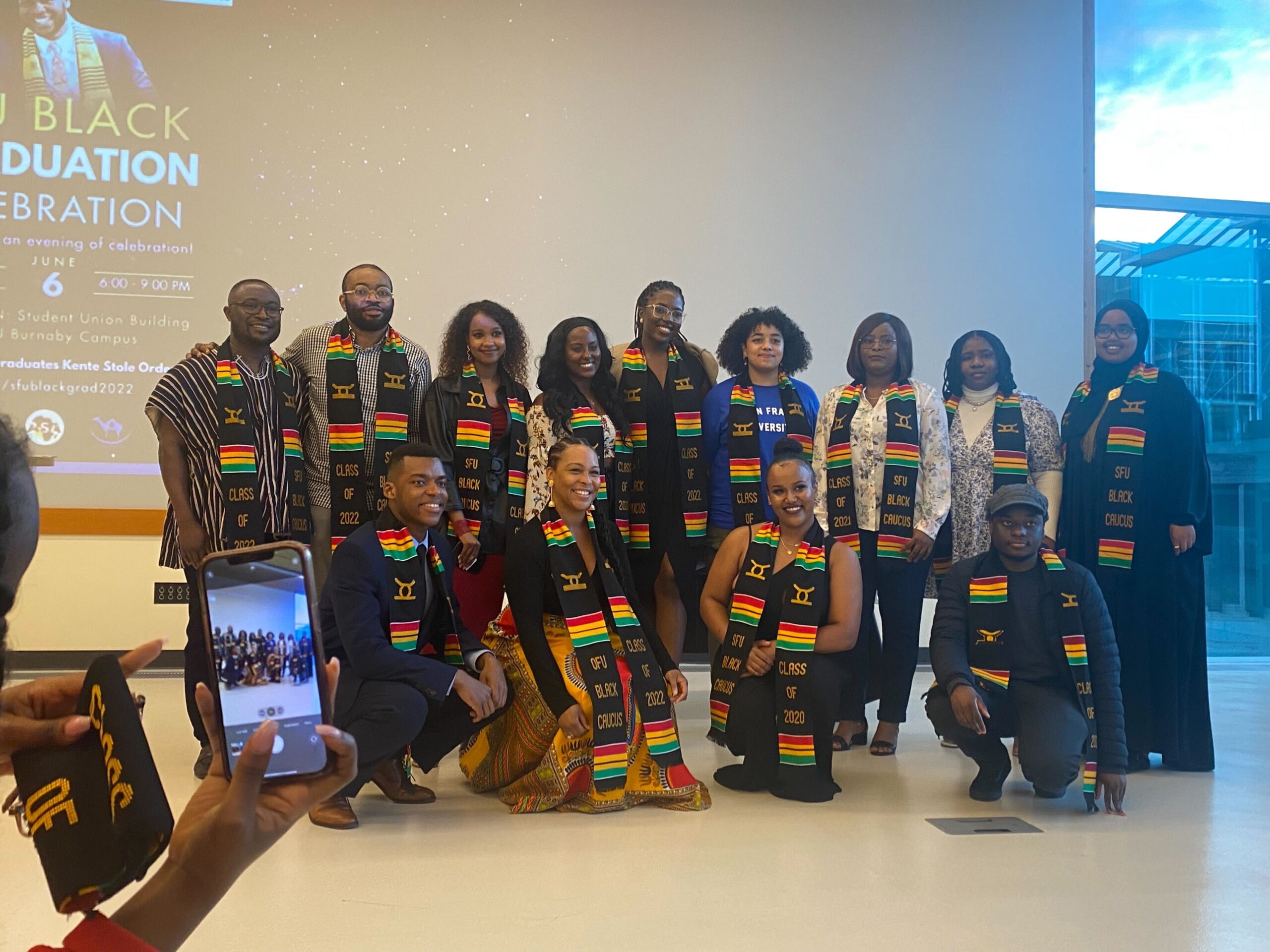 The photo shows 13 graduates wearing the Kente cloth stoles. They are huddled together smiling at the camera.