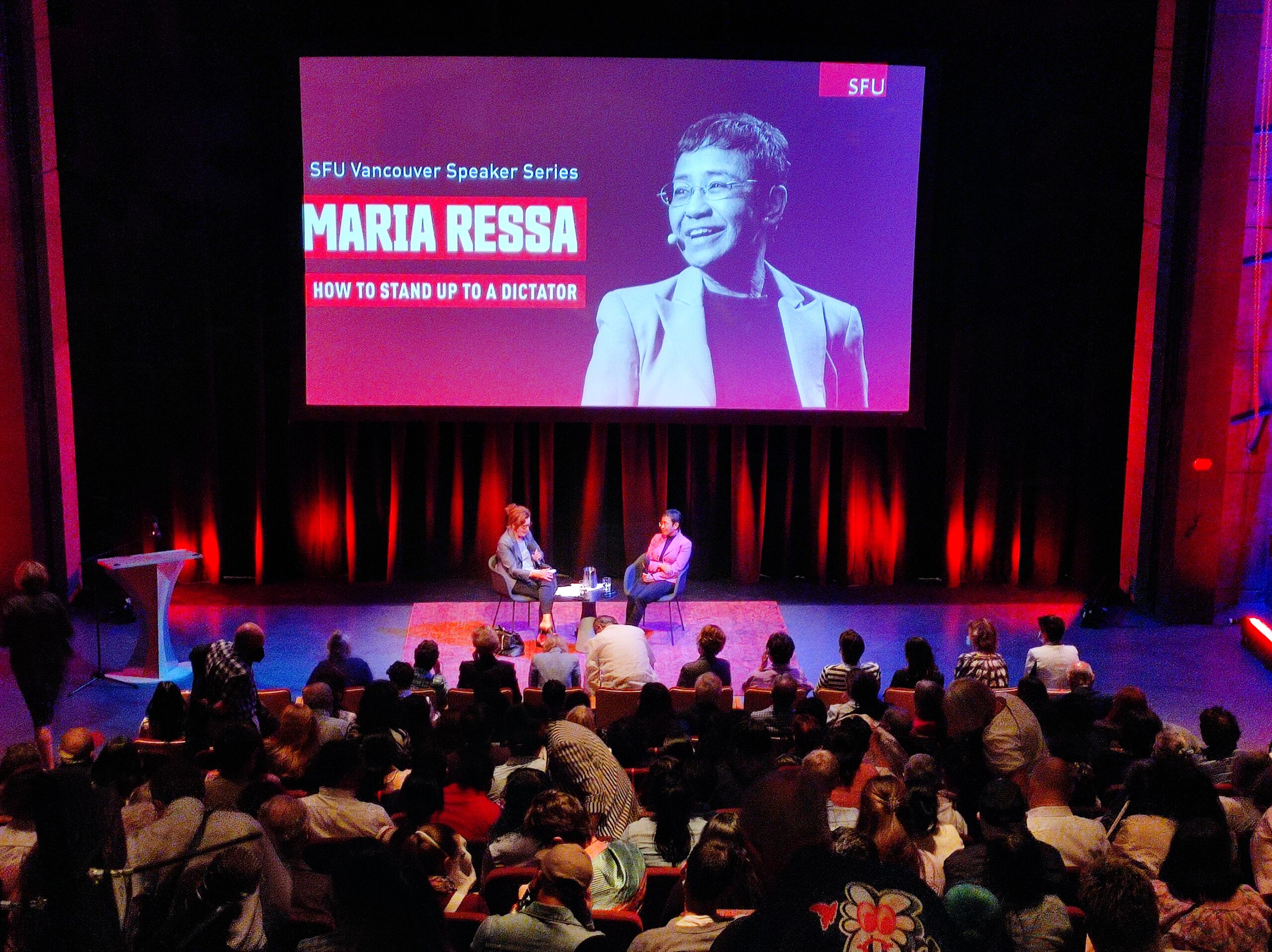 The photo is of Ressa and Off sitting on stage together as they engage in conversation. There are numerous people in the audience before them. A screen behind them has a large photo of Ressa and the title “How to Stand Up to a Dictator.”