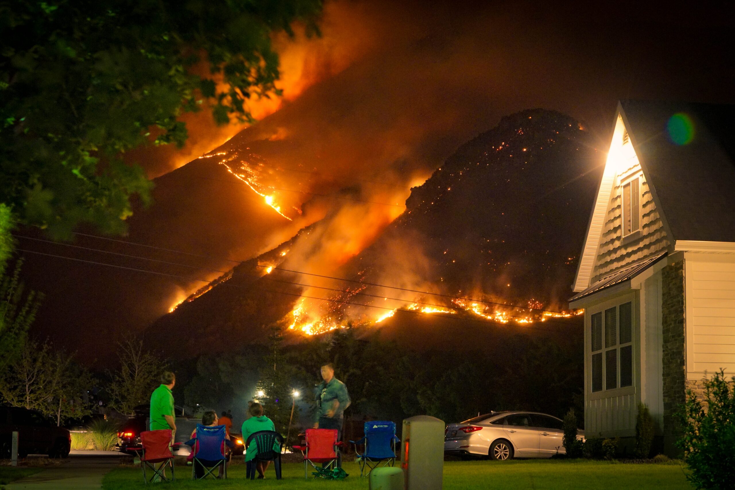 A group of people sit on a front lawn at night, directly overlooking a massive forest fire at night.