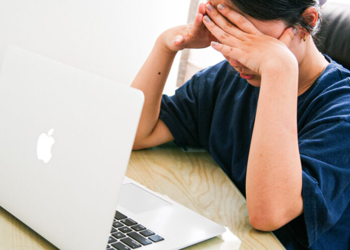 The photo is of a person sitting in front of their computer. Their head is in their hands and they look tired and stressed.