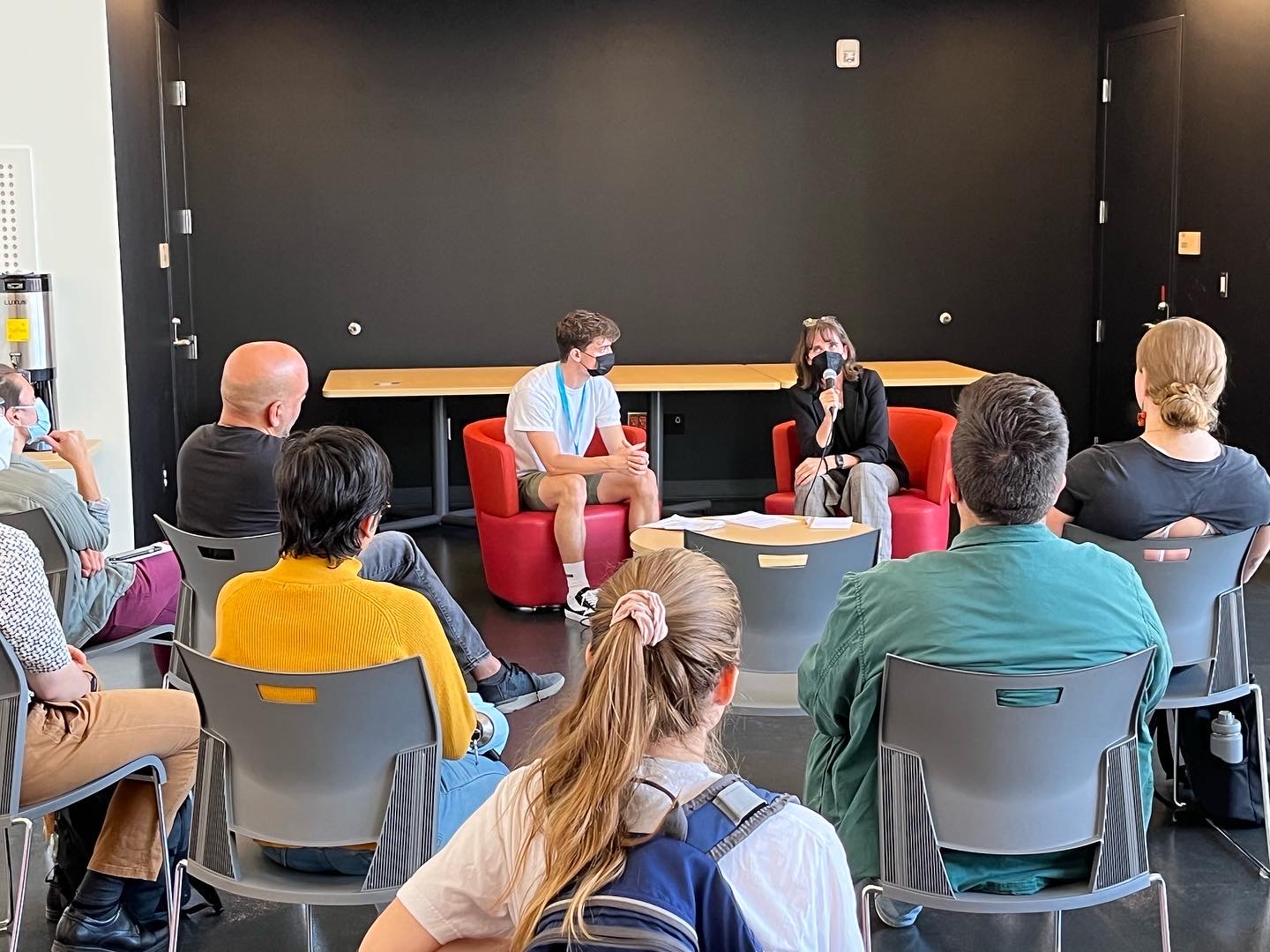 This photo is of Sonia Furstenau and Liam Mackay sitting at the front of an audience. They are engaged in conversation, sitting in two red chairs as people listen from the audience.