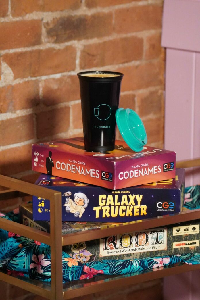 Stack of board games, with Code Names at the top, with a Mugshare mug on top filled with coffee. The board games are on a blue Hawaiian chair cushion behind a brick wall.