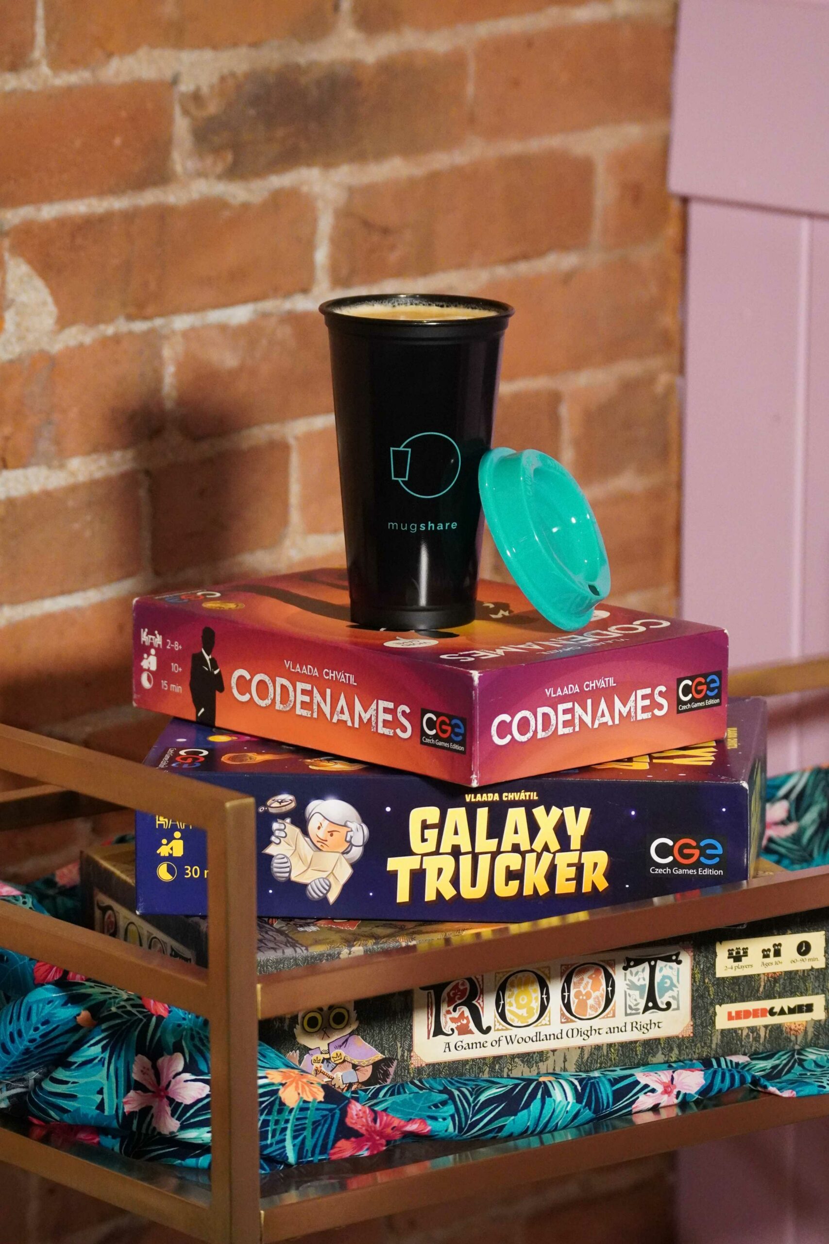 Stack of board games, with Code Names at the top, with a Mugshare mug on top filled with coffee. The board games are on a blue Hawaiian chair cushion behind a brick wall.