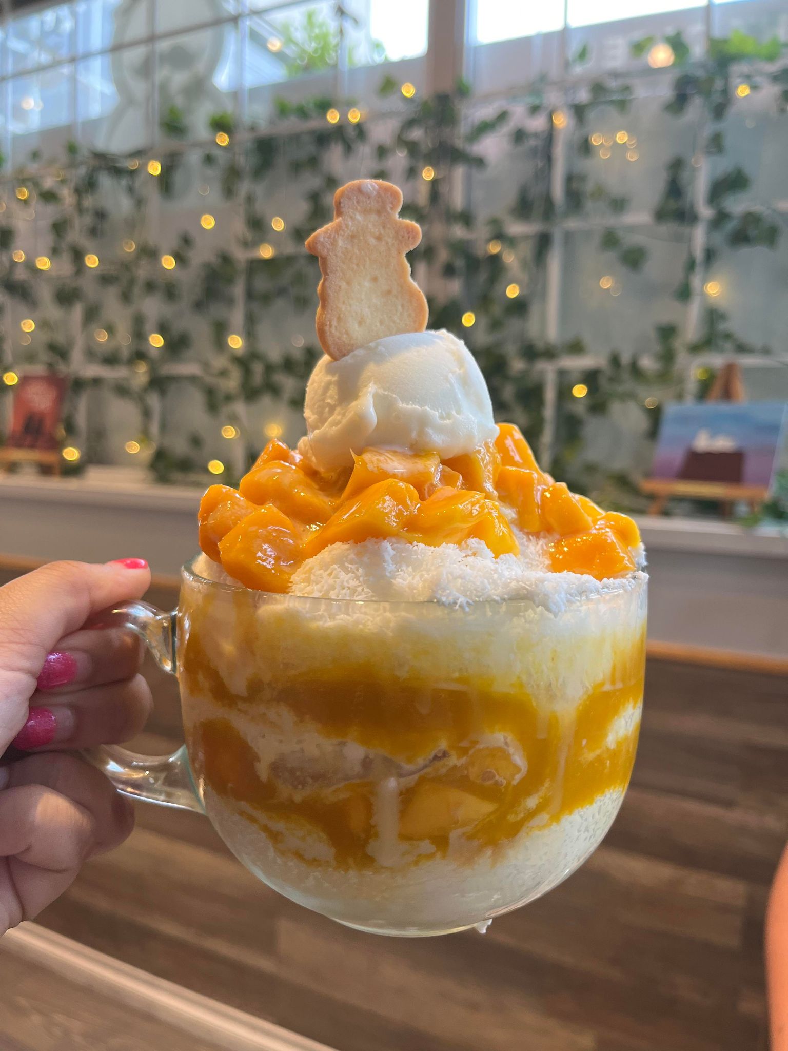 A clear glass of shaved ice topped with mango, a scoop of ice cream, and a cookie held up in front of a wall with greenery and string lights.