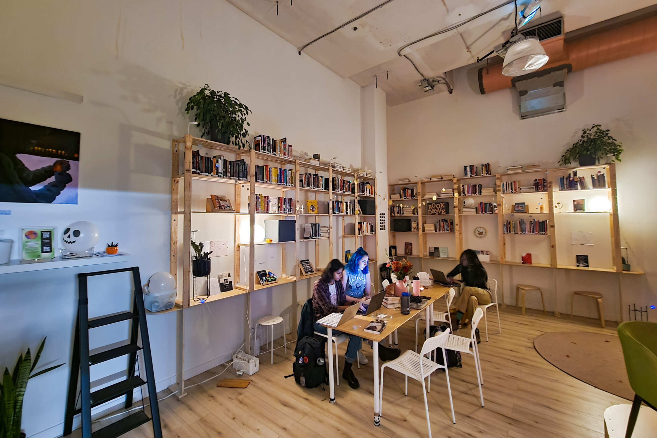 the interior of Vancouver Black Library: People working with their laptops on a table in the middle of a lofty room with book shelves against the walls, plants, and cozy lighting