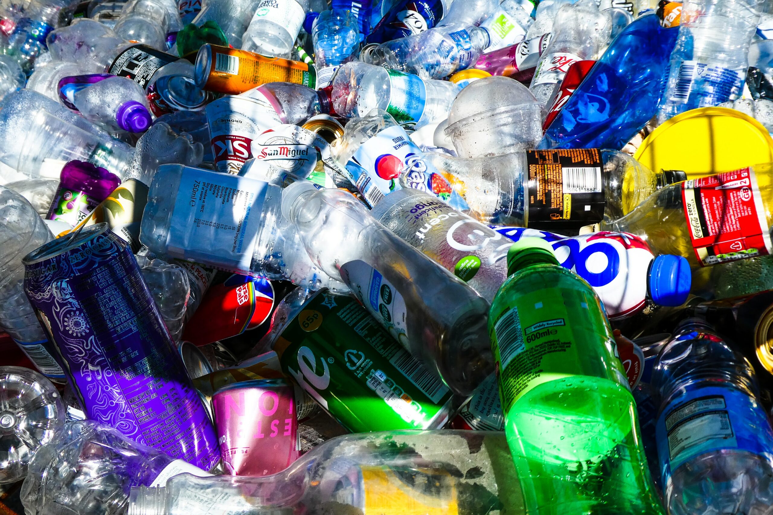 This photo is a close-up on recyclables. There are pop cans, plastic water bottles, and plastic containers piled on top of one another.