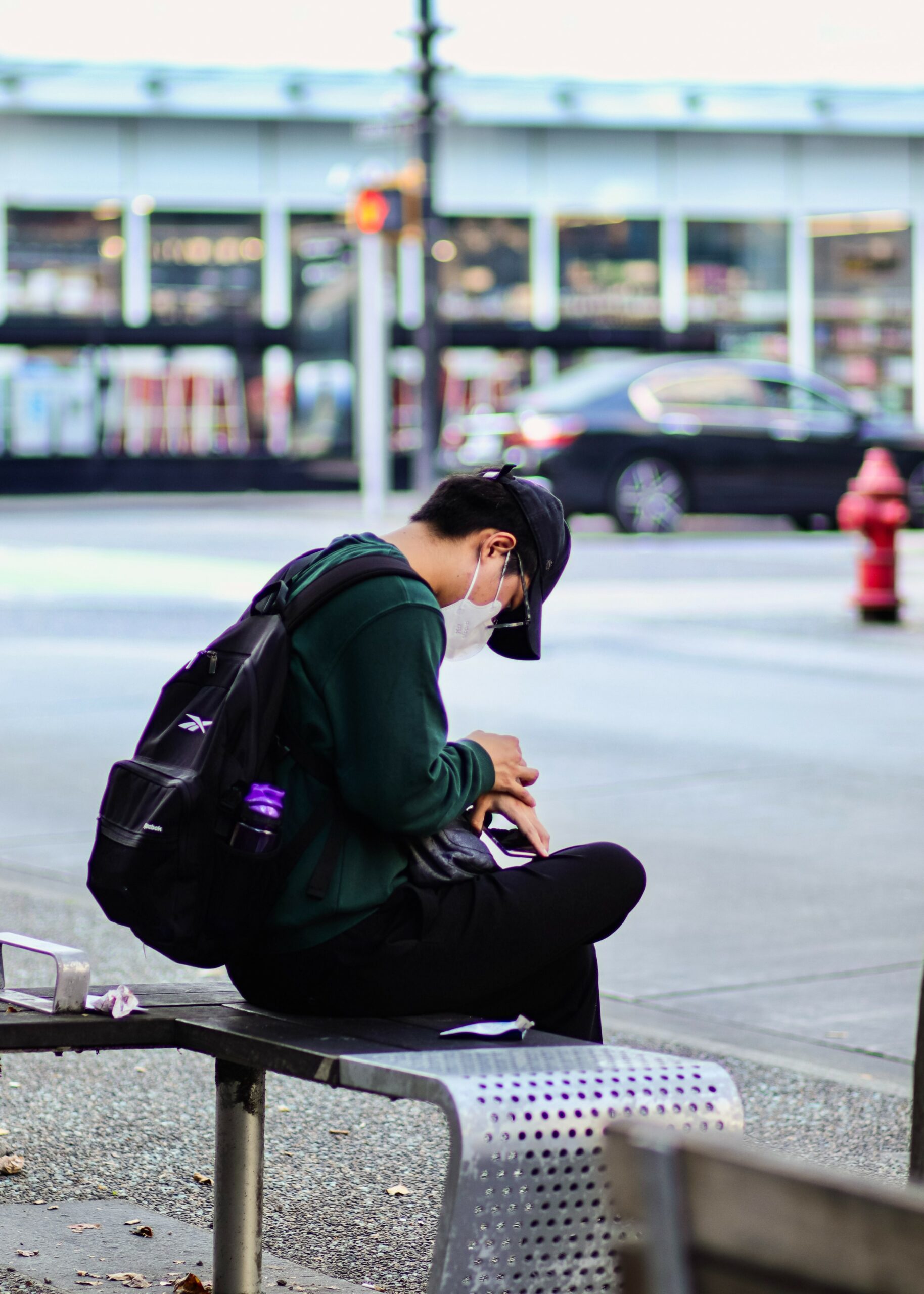 This is a photo of an individual sitting on a bench outside. They are wearing a mask while they look at their phone. They are sitting alone.
