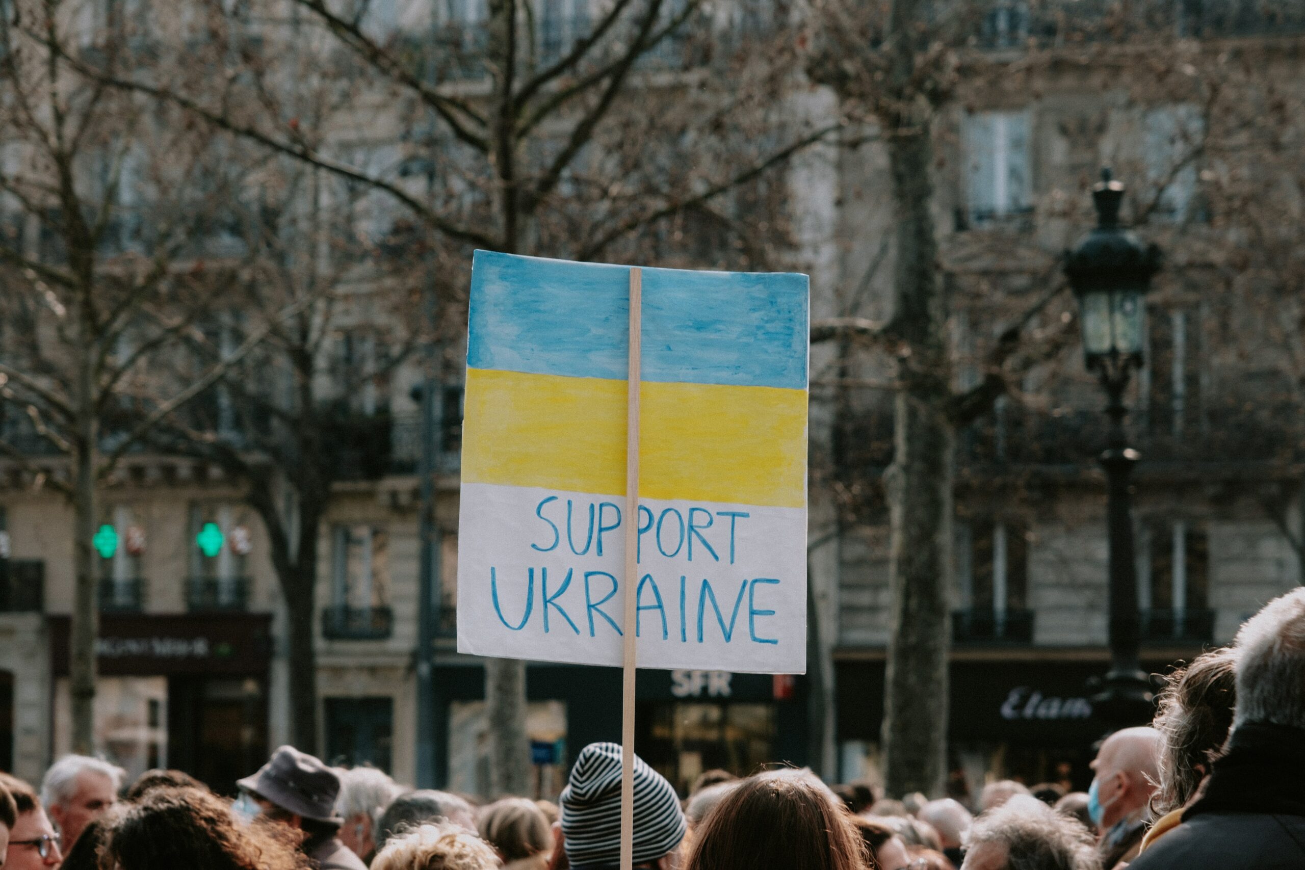 This photo is of a group of protestors. One individual is central in the photo and is holding a sign that reads “Support Ukraine.”