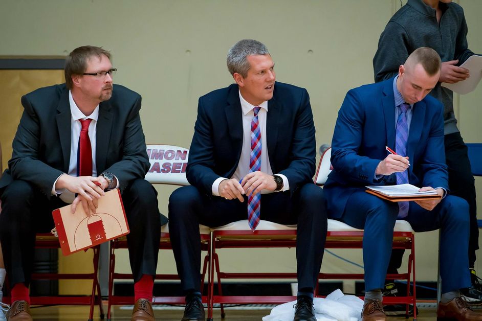 Photo of Steve Hanson and the basketball coaching staff sitting on the sideline during a game.