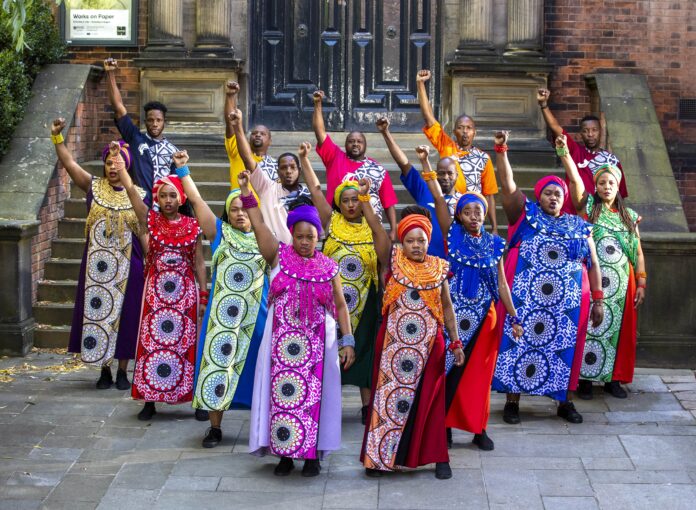 19 SWG Photo by Stephen Garnett-min Members of Soweto Gospel Choir in colorful traditional dress stand with their fists in the air.