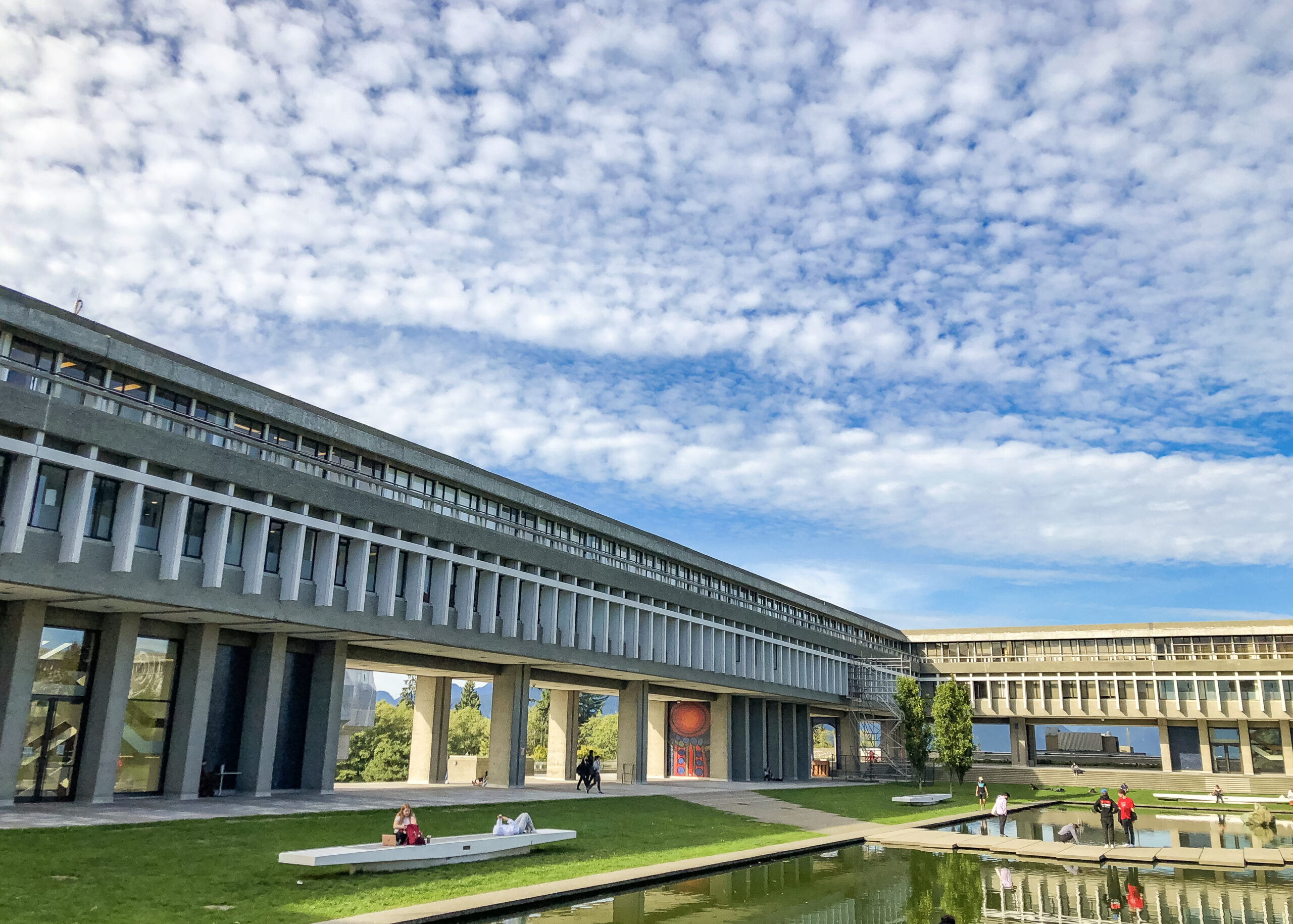 This is a photo of the academic quadrangle at the SFU Burnaby campus. In front of the building is students sitting on the green grass.