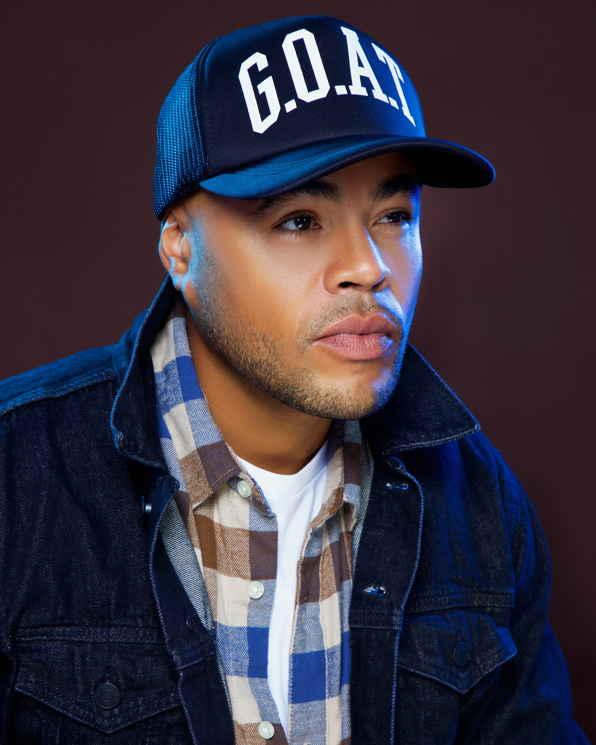 Headshot of D. O. Gibson. Gibson wears a blue hat that says “G.O.A.T.” and a blue plaid shirt against a black backdrop.