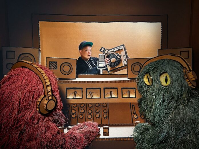 Kid Koala Photo of Kid Koala making music with a turntable and a cardboard cut-out of a recording studio. Two bright-colored monsters with headphones sit outside the recording room, operating the recording settings.
