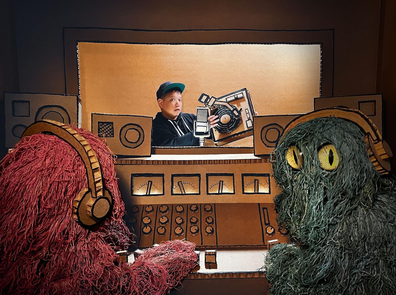 Photo of Kid Koala making music with a turntable and a cardboard cut-out of a recording studio. Two bright-colored monsters with headphones sit outside the recording room, operating the recording settings.