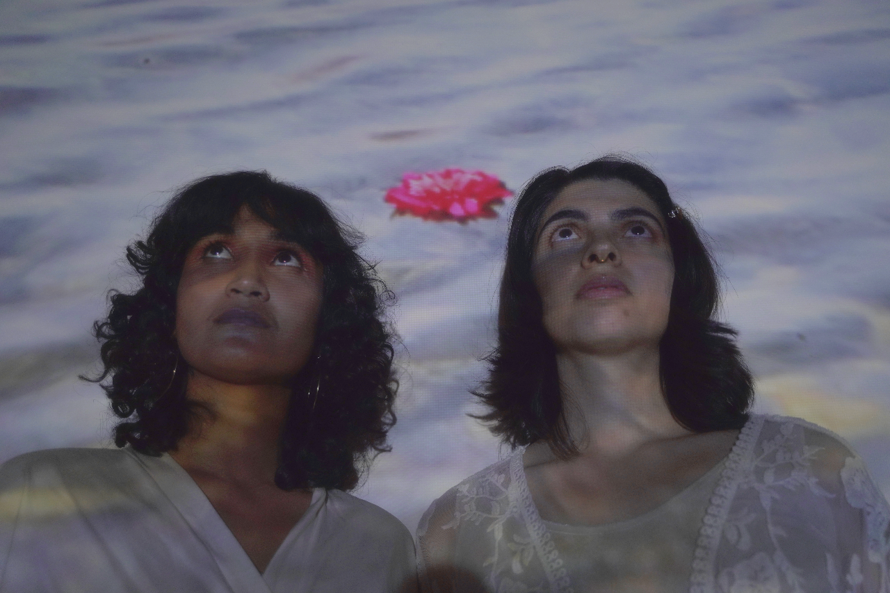 Anjelica Solomon and Oceaan Pendharkar in white blouses look up with opaque, shimmering water and a pink lily pad in the background.