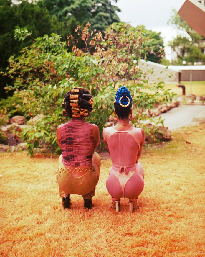 Two Black femme-presenting people are crouched in what looks like a grassy front yard, with only their backsides visible. Both have dynamic disco-esque outfits on, one with hair rollers holding up their hair, and the other with a top knot and traditional hat.