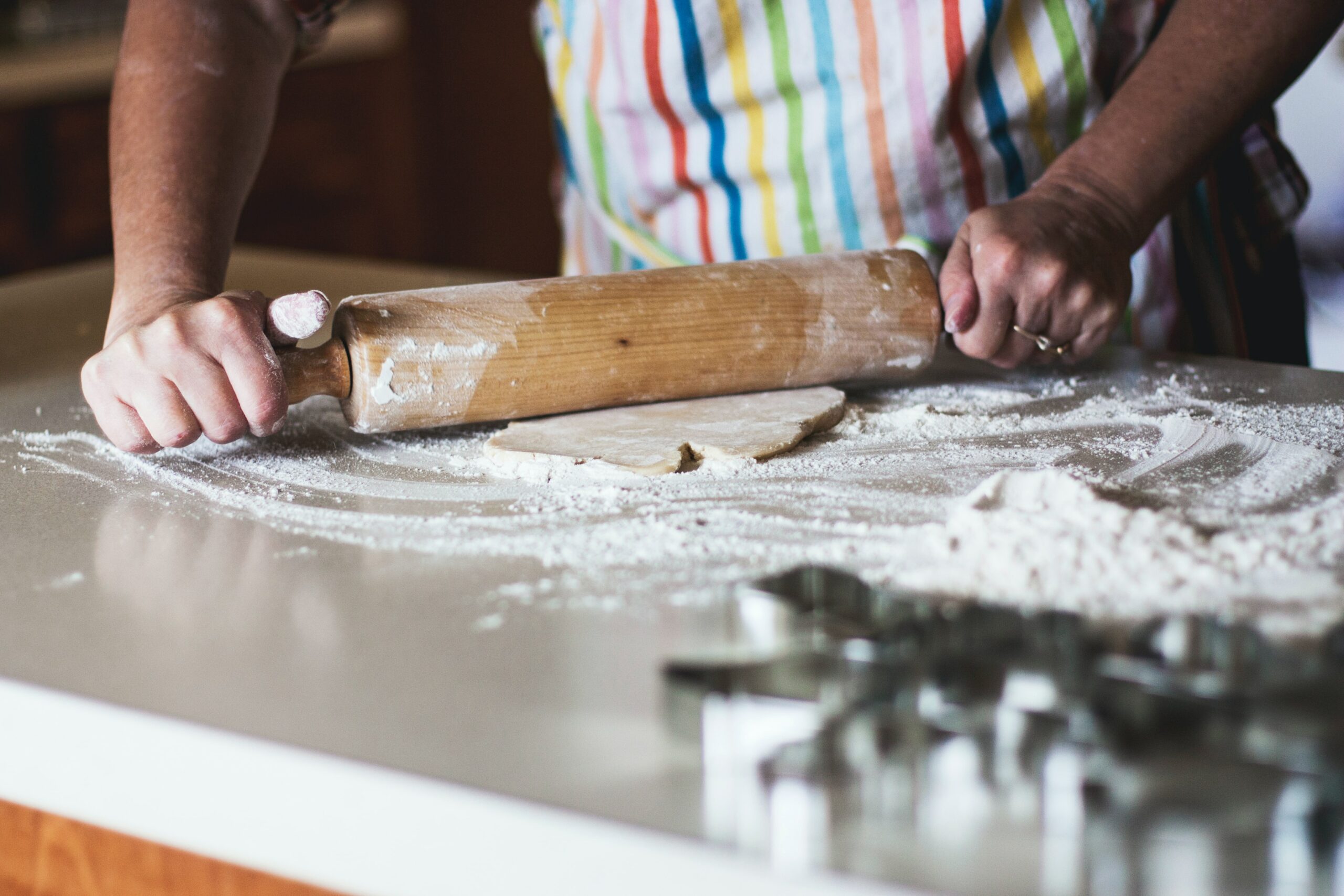 Person pictured arms down wearing a rainbow-striped apron rolling out dough on a floured kitchen counter.