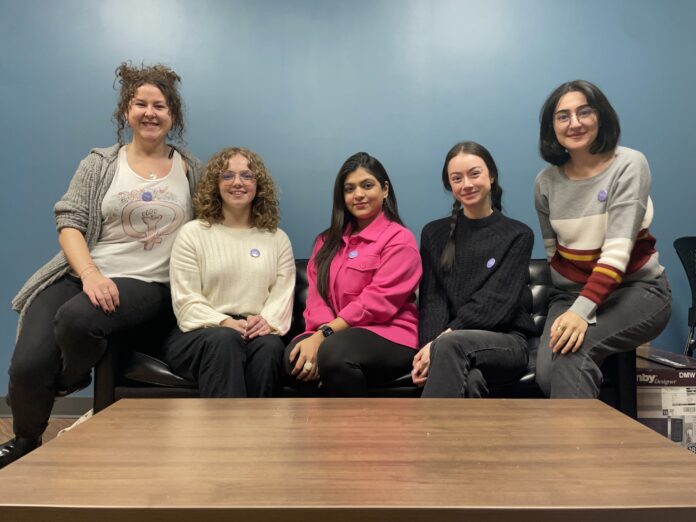 This is a photo of five of the organizers of the event. They are sitting on a couch smiling at the camera and behind them is a blank blue wall