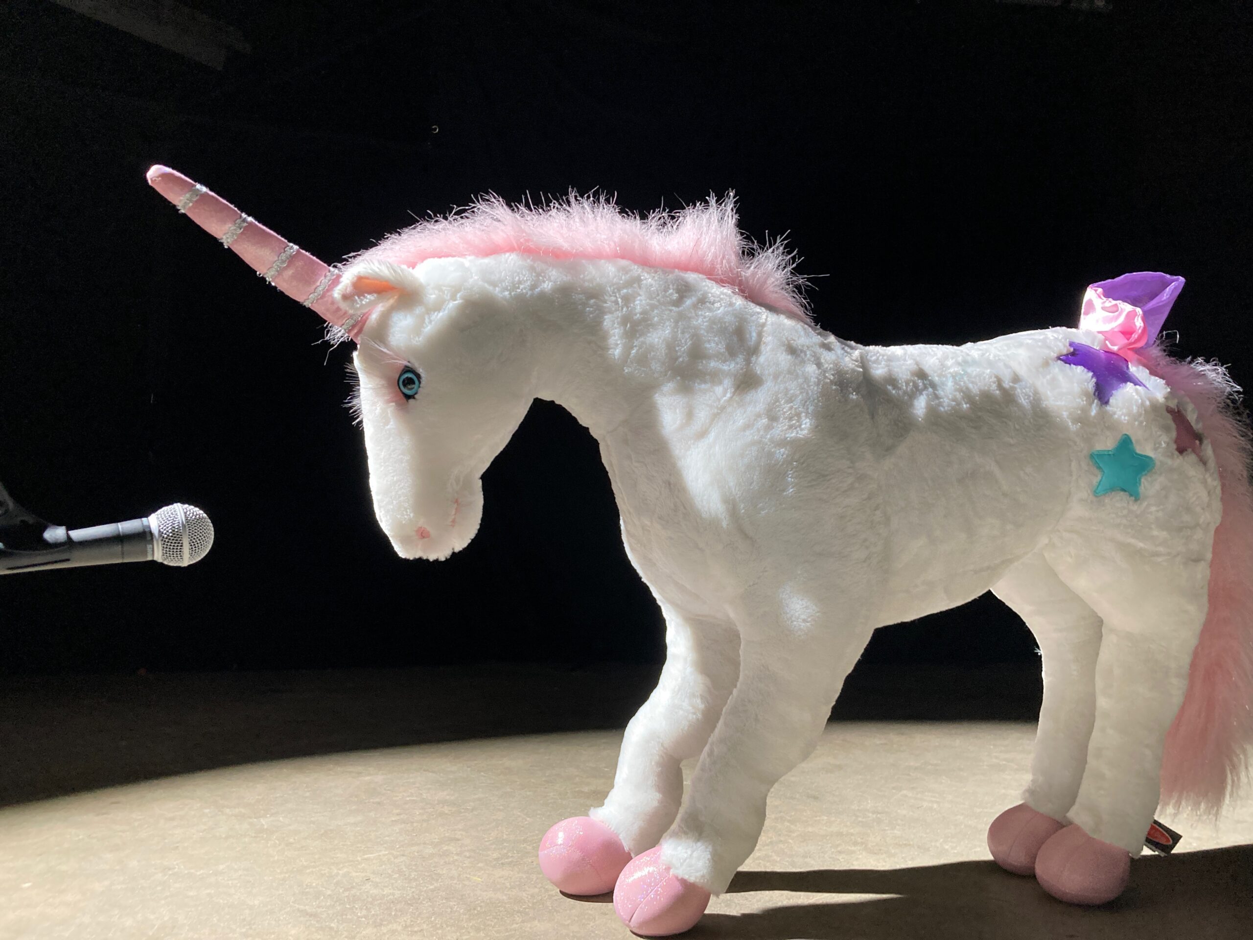 A stuffed unicorn on a stage standing in front of a mic.