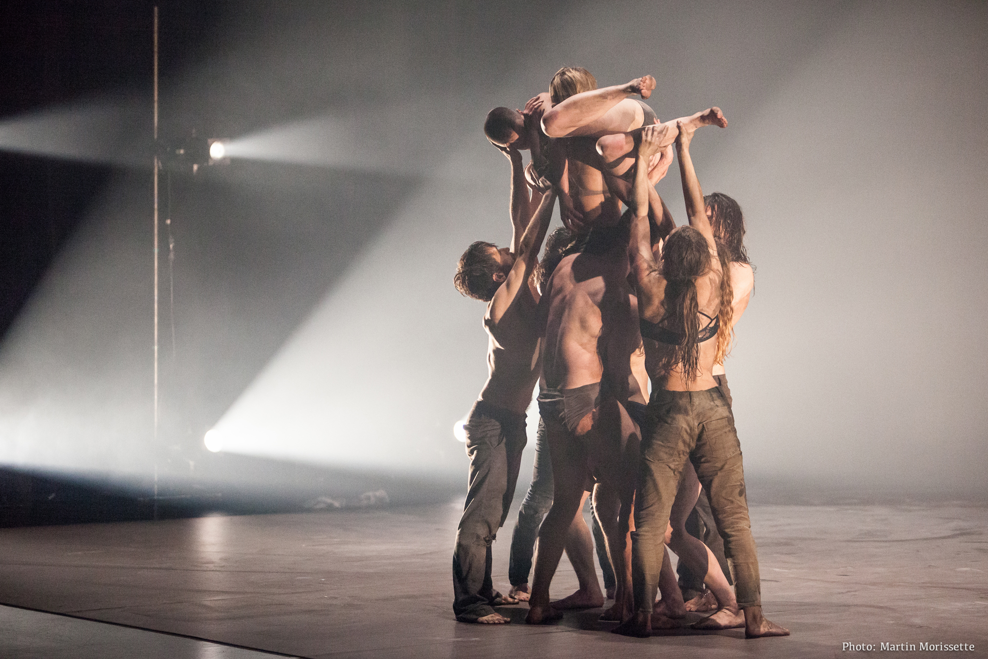 A group of shirtless dancers with black pants lift two dancers up.