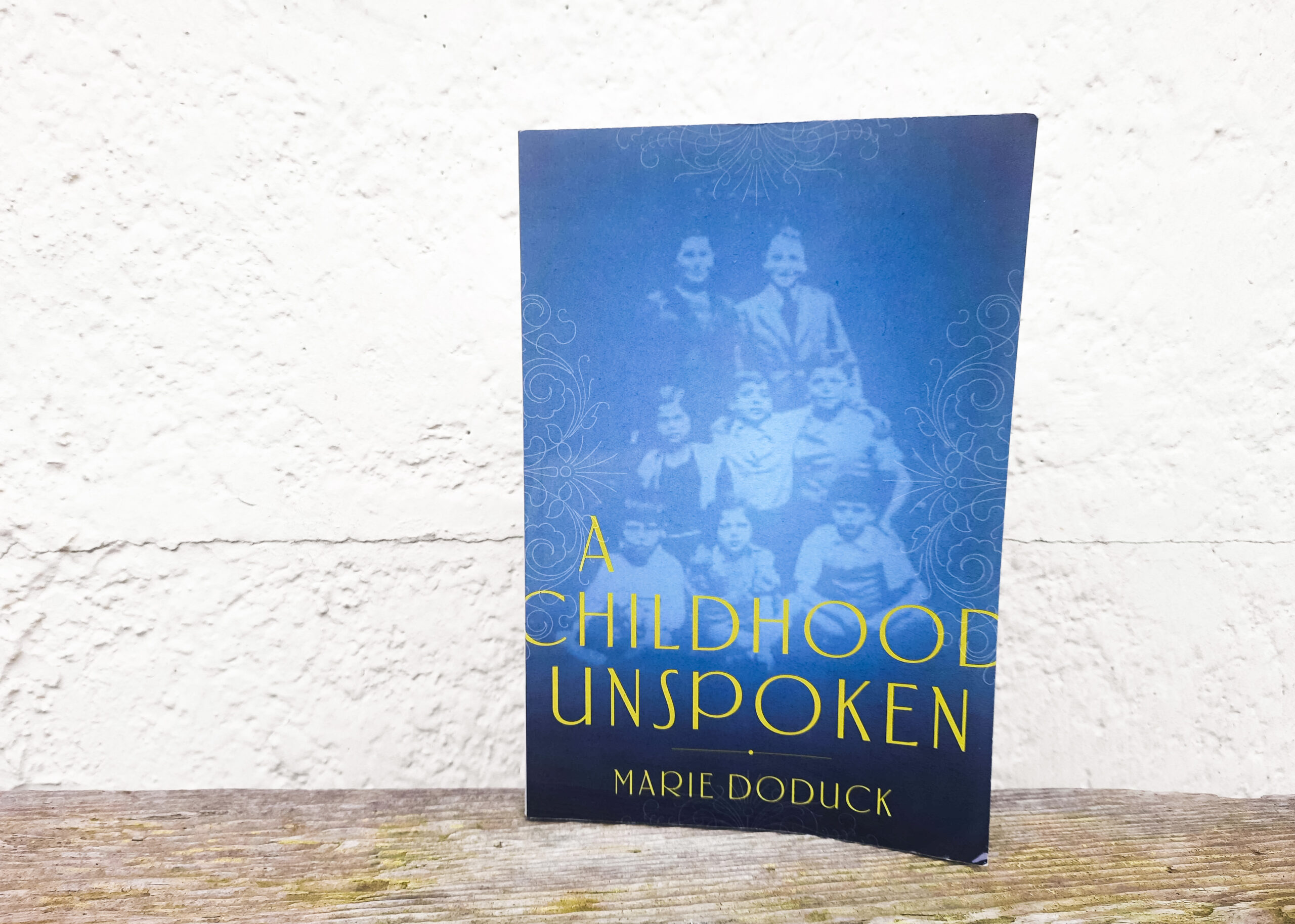 This is a photo of the front cover of Marie Doduck’s memoir. The cover is blue and has a faint photo of Marie Doduck’s siblings as children.