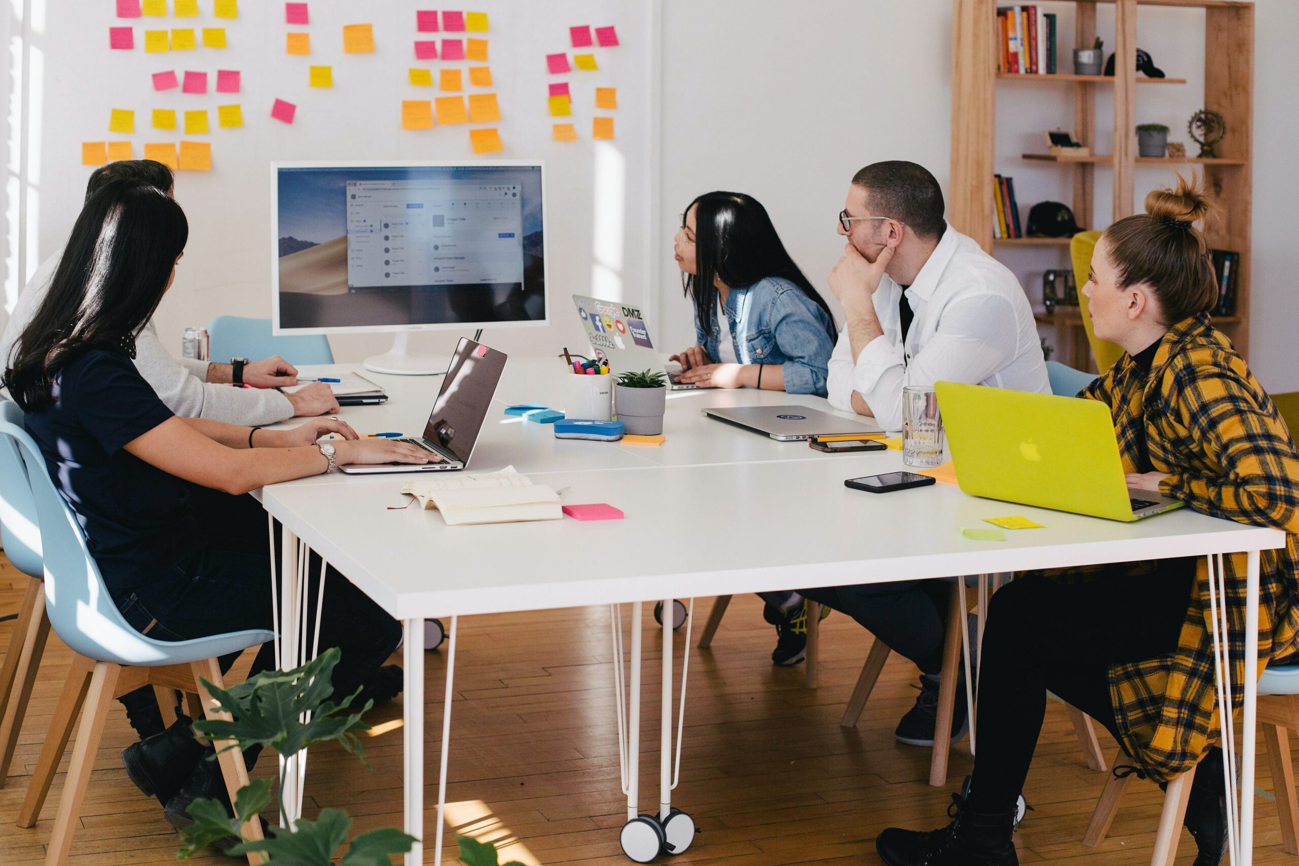 This is a photo of a group of young people collaborating in an office. They have laptops in front of them but they are looking at a TV at the front of the room.