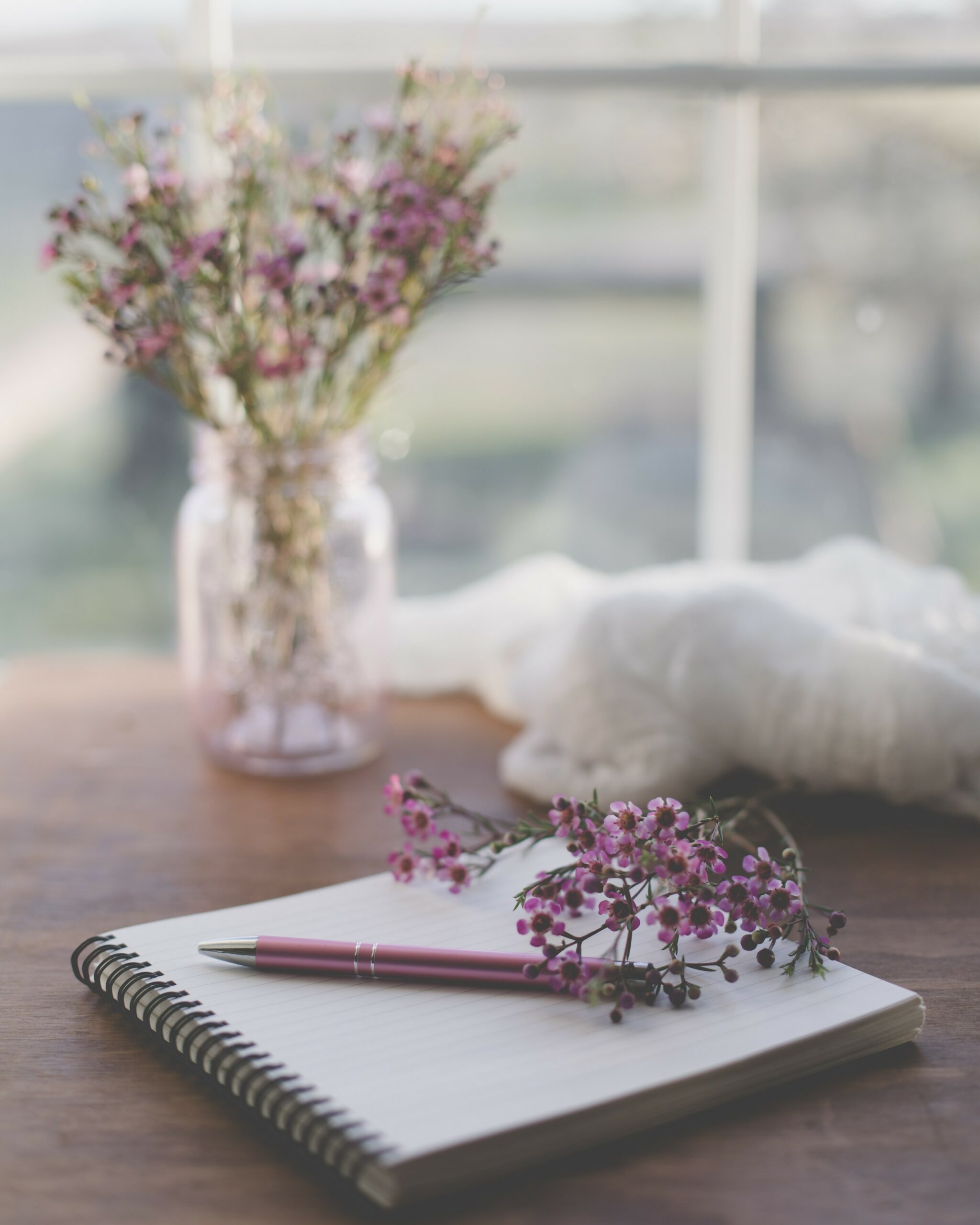 A lined notebook on a wooden table with a pen and purple flowers on top.