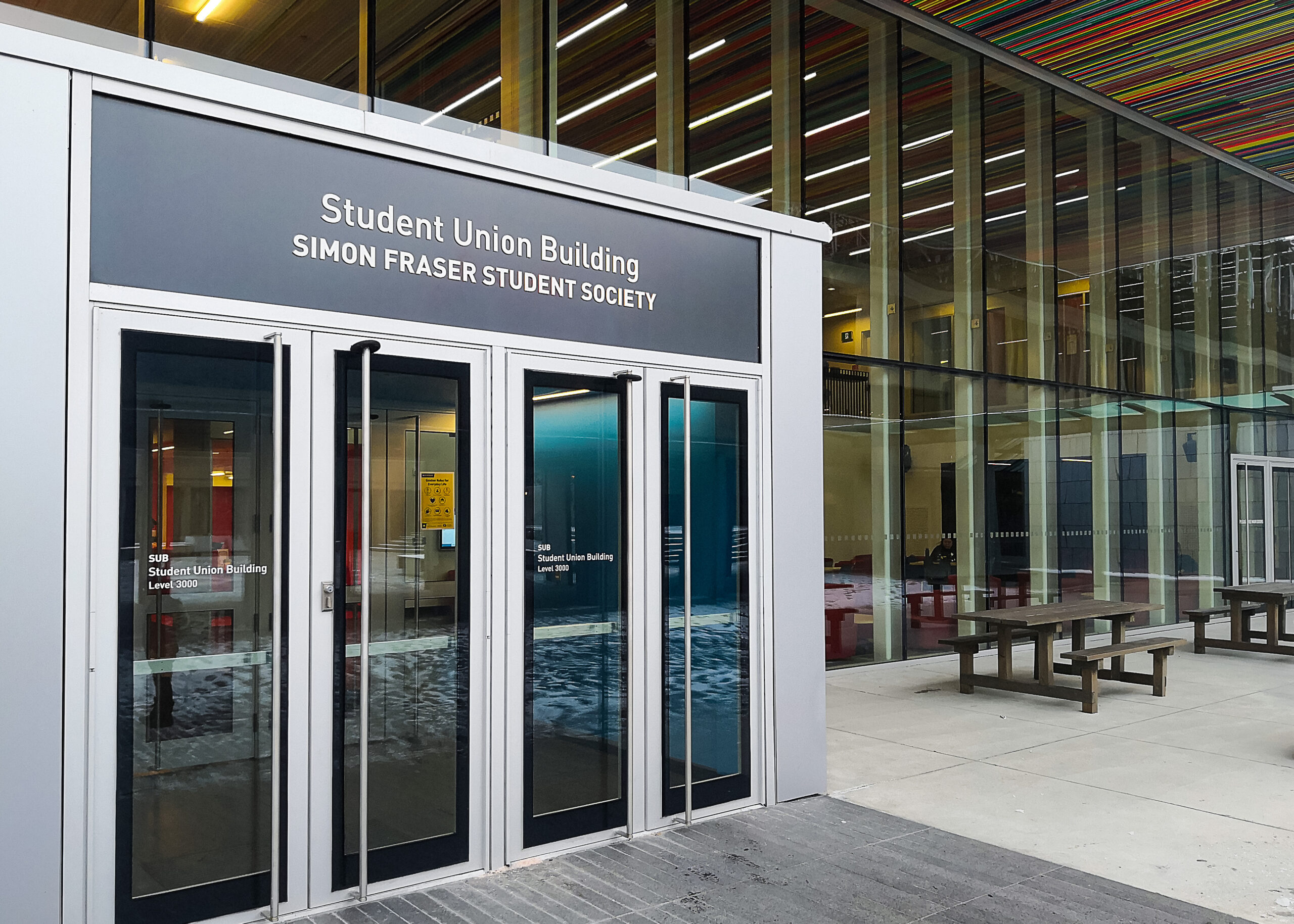 This is a photo of the outside of the Student Union Building at the SFSS Burnaby campus. The photo is a close up of the doors. Above the doors the sign reads “Student Union Building, Simon Fraser University”