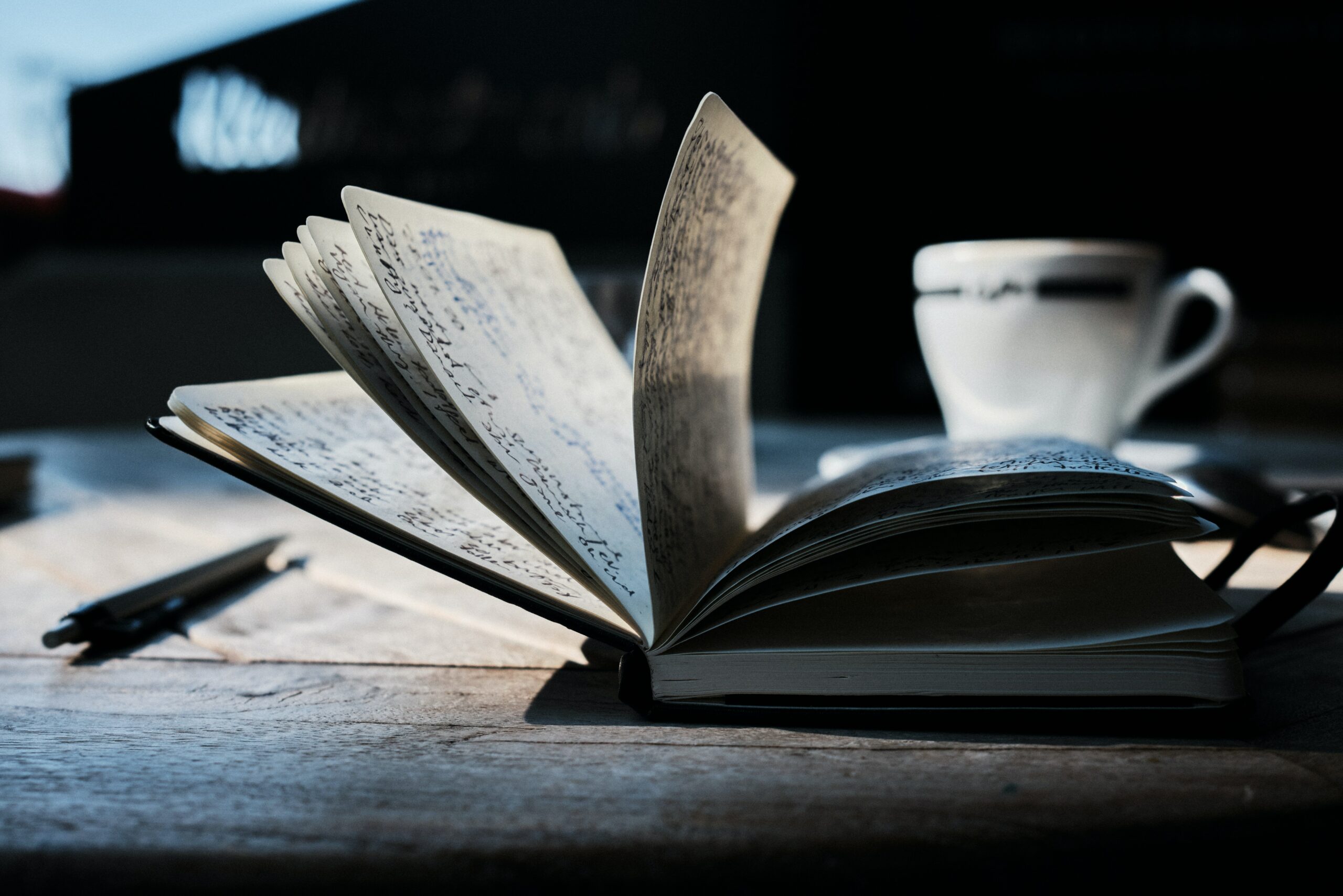 An open notebook with many pages opened to show cursive writing in black ink on the pages. A pen and a tea cup are in the background.