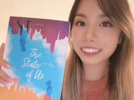 Elvira Chan holds her book, displaying the cover with an illustration of two shadows of people walking against a backdrop of tall buildings on each side of the book. Between them is a watercolor gradient of a blue, cloudy sky. They are each on opposite sides of the book— one on top and one on the bottom. The one on top is upside down. Chan is smiling wearing an orange knit sweater.