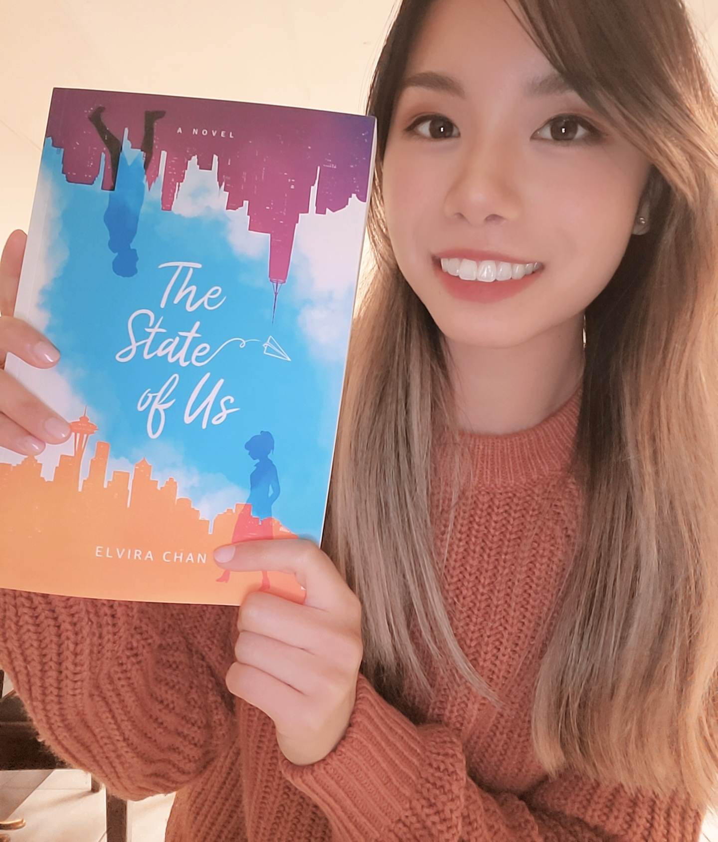 Elvira Chan holds her book, displaying the cover with an illustration of two shadows of people walking against a backdrop of tall buildings on each side of the book. Between them is a watercolor gradient of a blue, cloudy sky. They are each on opposite sides of the book— one on top and one on the bottom. The one on top is upside down. Chan is smiling wearing an orange knit sweater.