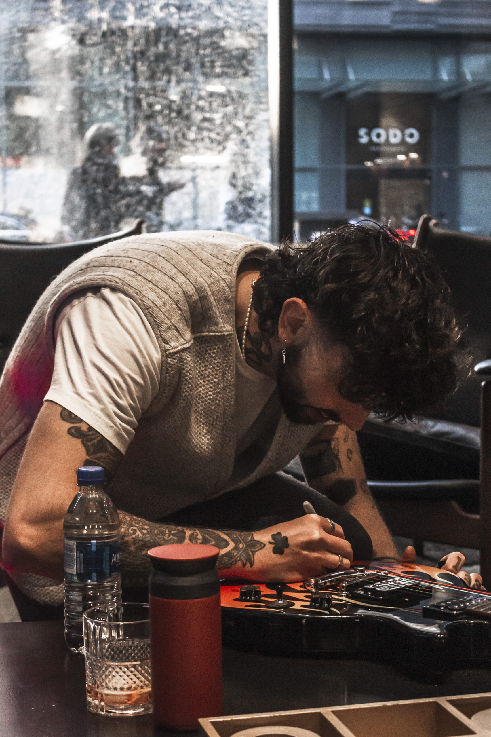 Emét is seated and signing a guitar. They are wearing a gray sweater vest on top of a white T-shirt with tattoos on their arms. Behind them, there is a window that shows snow outside.