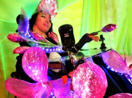 An Indigenous model in a wheelchair wearing a shimmery dress made of fireweed flower. The dress consists of large fireweed flowers and flower petals adorning the wheelchair, with LED lights and purple fringe.