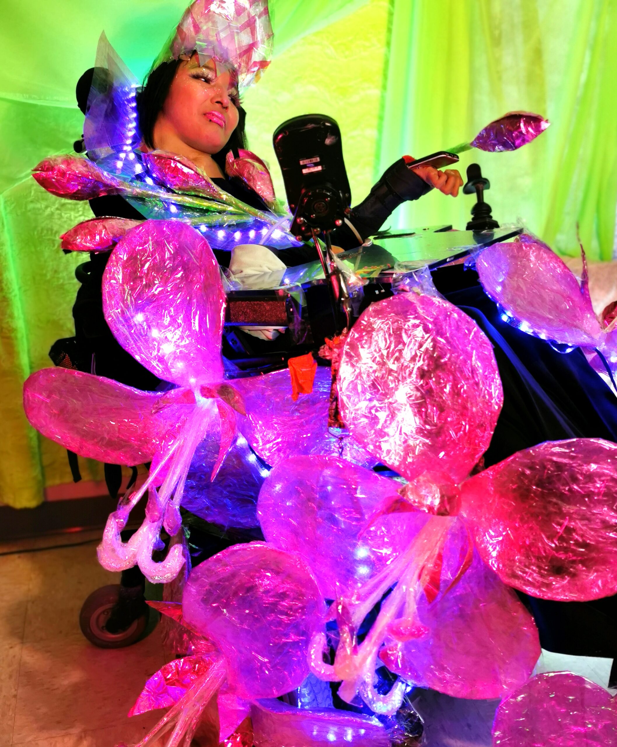 An Indigenous model in a wheelchair wearing a shimmery dress made of fireweed flower. The dress consists of large fireweed flowers and flower petals adorning the wheelchair, with LED lights and purple fringe.