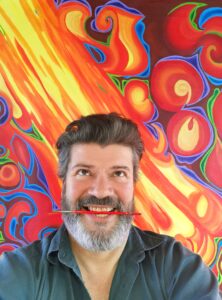 Michel poses for a selfie with a joyful expression on his face and a paintbrush between his teeth. He is standing in front of a fiery painting with vibrant yellows and reds with a touch of blue swirls.