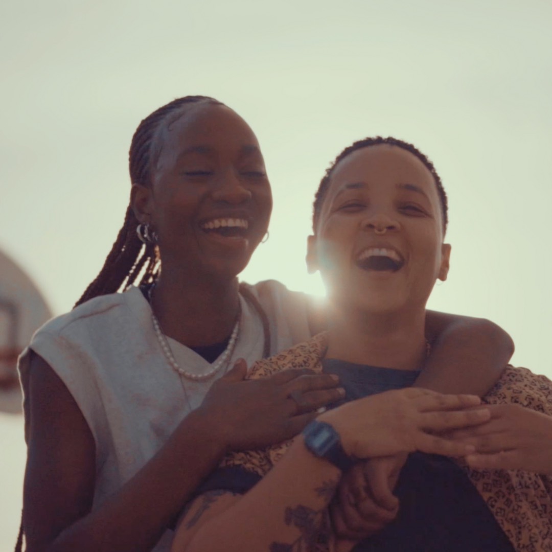 A woman with a pearl necklace, hoop earrings, and cornrow braids wraps her arms around OK Naledi, who is wearing a tan collar shirt. Both of them are smiling with their mouths wide. A basketball hoop is seen blurred behind them, and the rest of the background is a gray-ish blue sky.