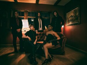 This is a photo of the five members of VERTTIGO looking fiercely to the camera. They are all wearing black clothes. Two members sit on a brown couch wearing leather and three members stand behind them. The image is a shadowy interior of an antique-looking room with carpet, a painting on the wall with an antique frame, a window, and a light shining from behind them.