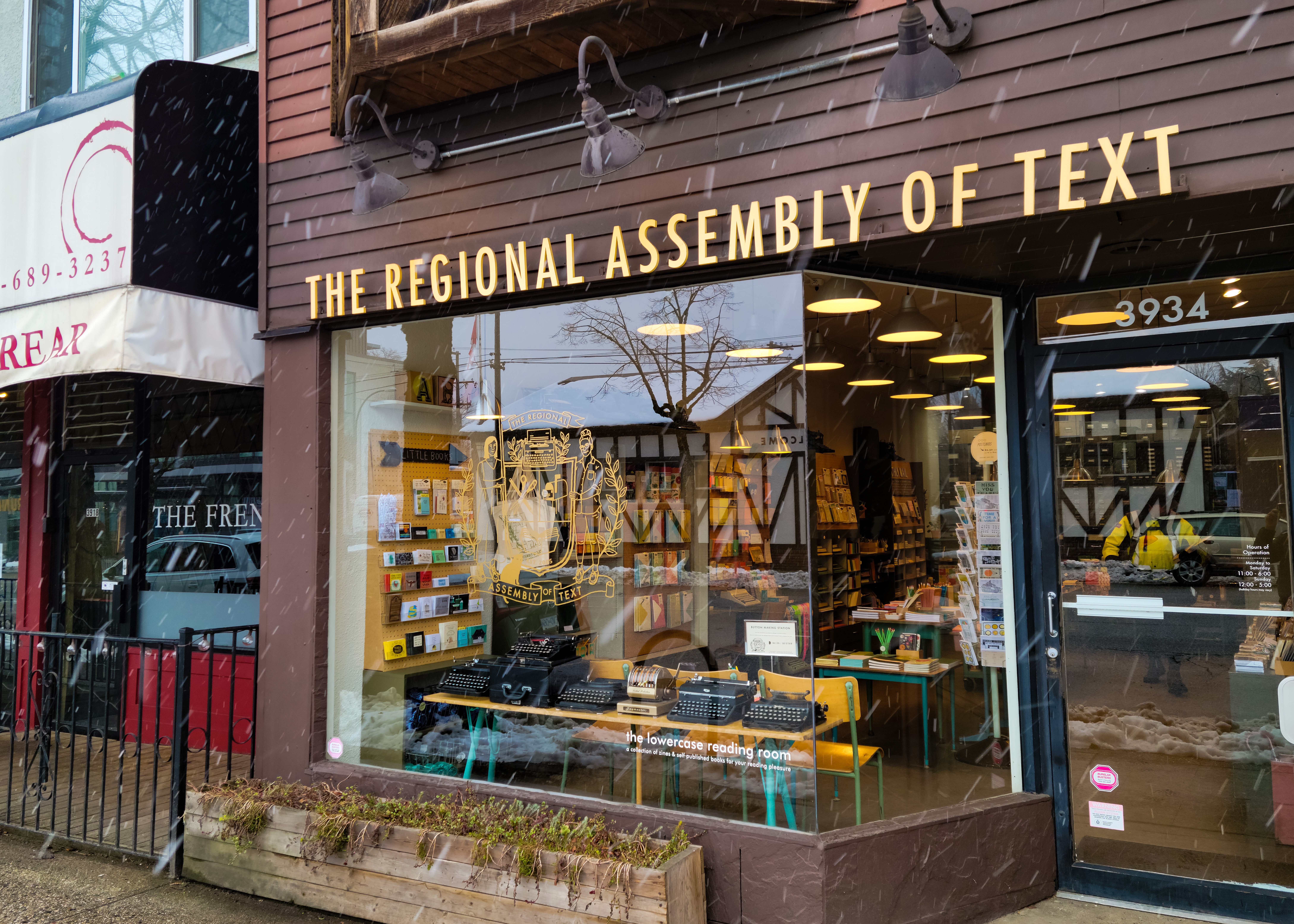 The exterior of a building with chestnut-coloured wooden panels and text sitting above a window that reads “Regional Assembly of Text.” There are snowflakes falling outside and trees reflected in the window. There are various typewriters in the display window.