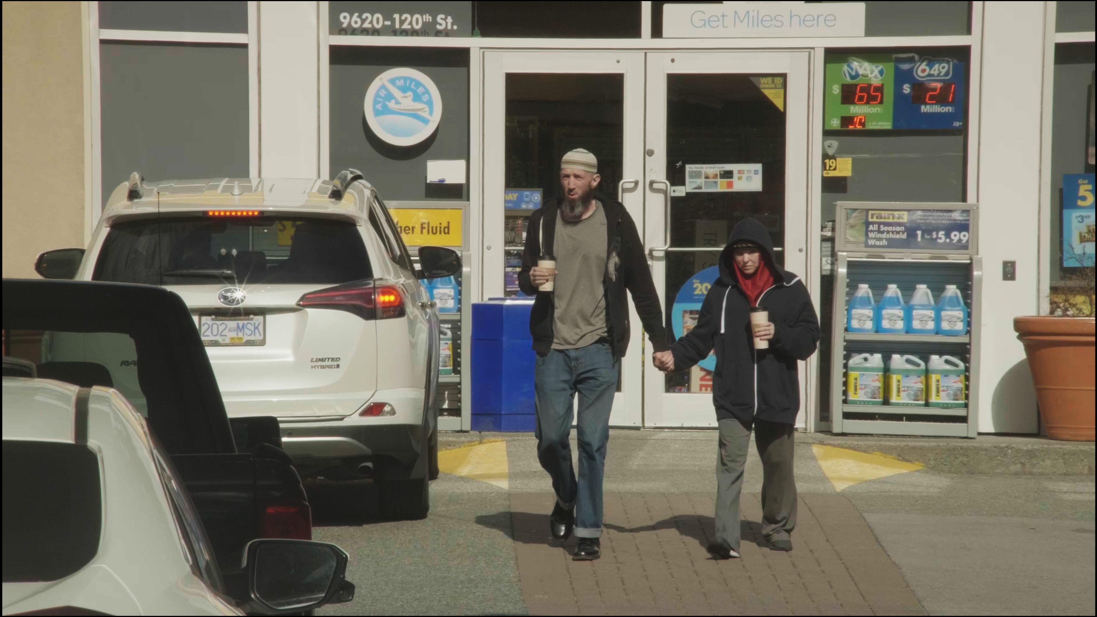 FILM STILL: Ana and Omar walk outside of a gas station convenience store holding hands and each have a paper cup. Ana is wears a black hoodie over a red hijab and baggy pants and Omar wears a white kufi on his head with brown stripes, a black jacket, jeans, and an earth-tone T-shirt.