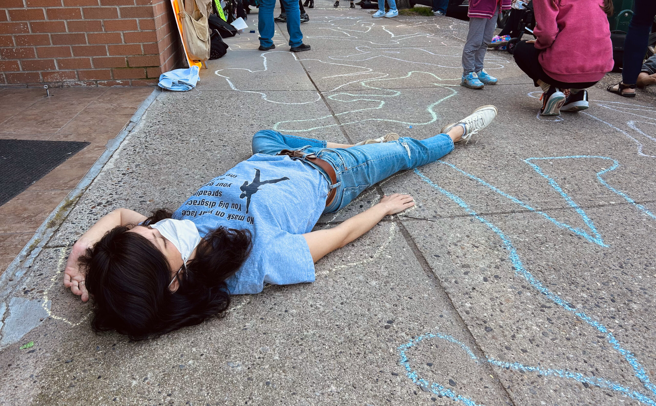 On the pavement in the foreground lies a young protester with shoulder-length brown hair, a blue t-shirt and jeans, and an N95 repirator mask. They lie as if dead, one hand fallen beside their face, surrounded by a chalk body outline. All around them are other chalk body outlines stretching as far as the eye can see. In the corners of image are the feet of standing and crouching protesters.