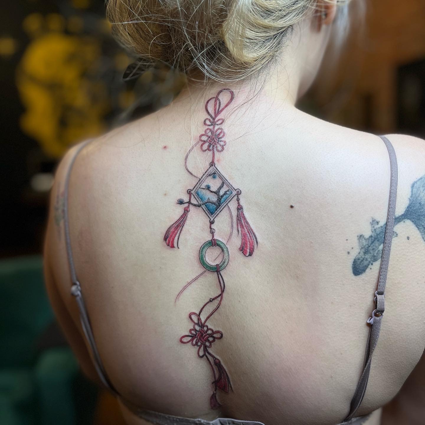 A photo of a coloured tattoo that flows from the back of their neck and down their spine. The tattoo is an East-Asian style charm made up of red string with tassels and beautiful floral knots. The string is tied to a jade ring, small beads, and a miniature framed photo of a tree with a branch emerging out of the photo behind a blue background.