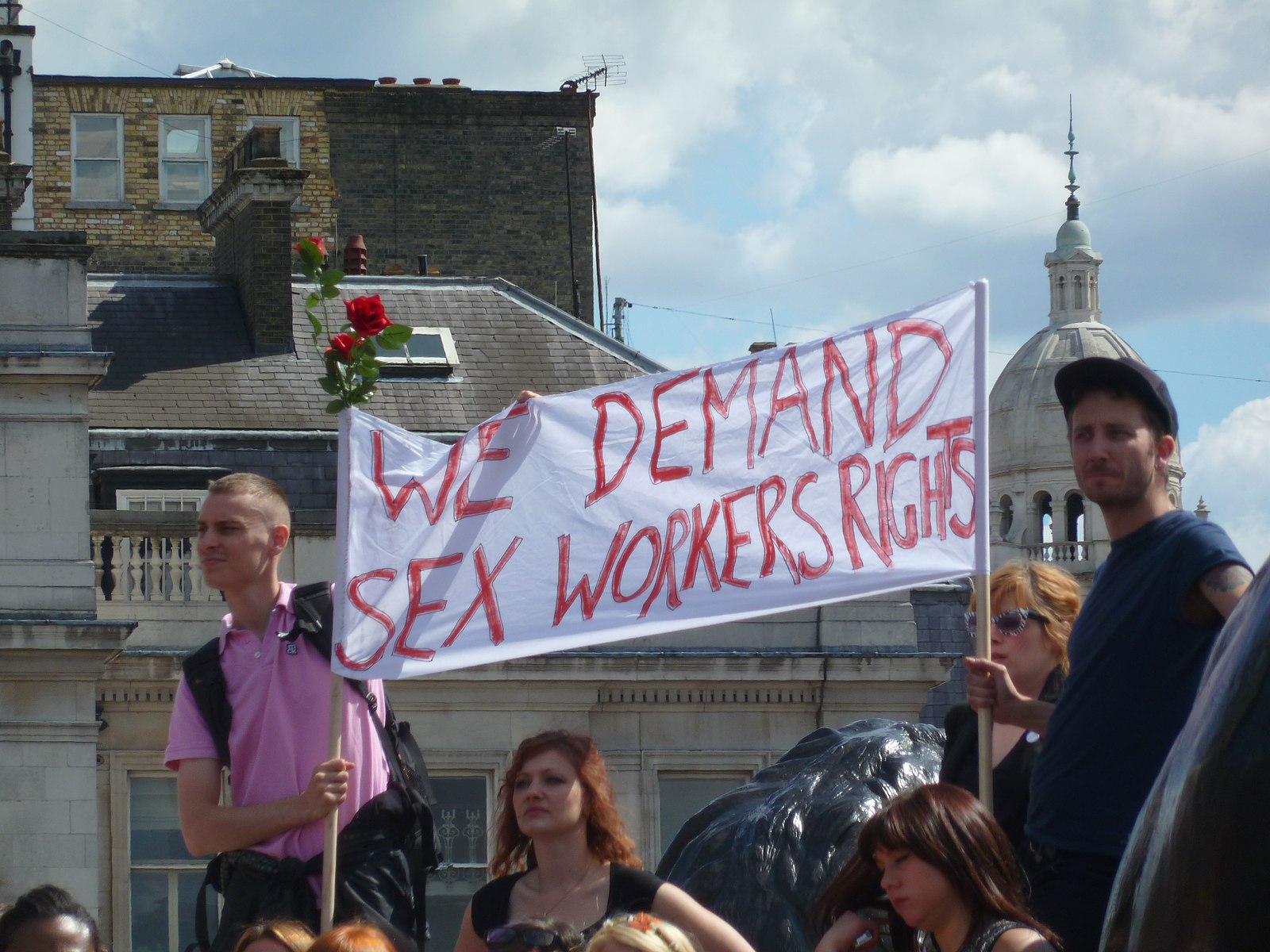 This is a photo from the 2011 Slut Walks in the United Kingdom. Protestors are holding a large banner that reads “We Demand Sex Worker’s Rights,” in red font.