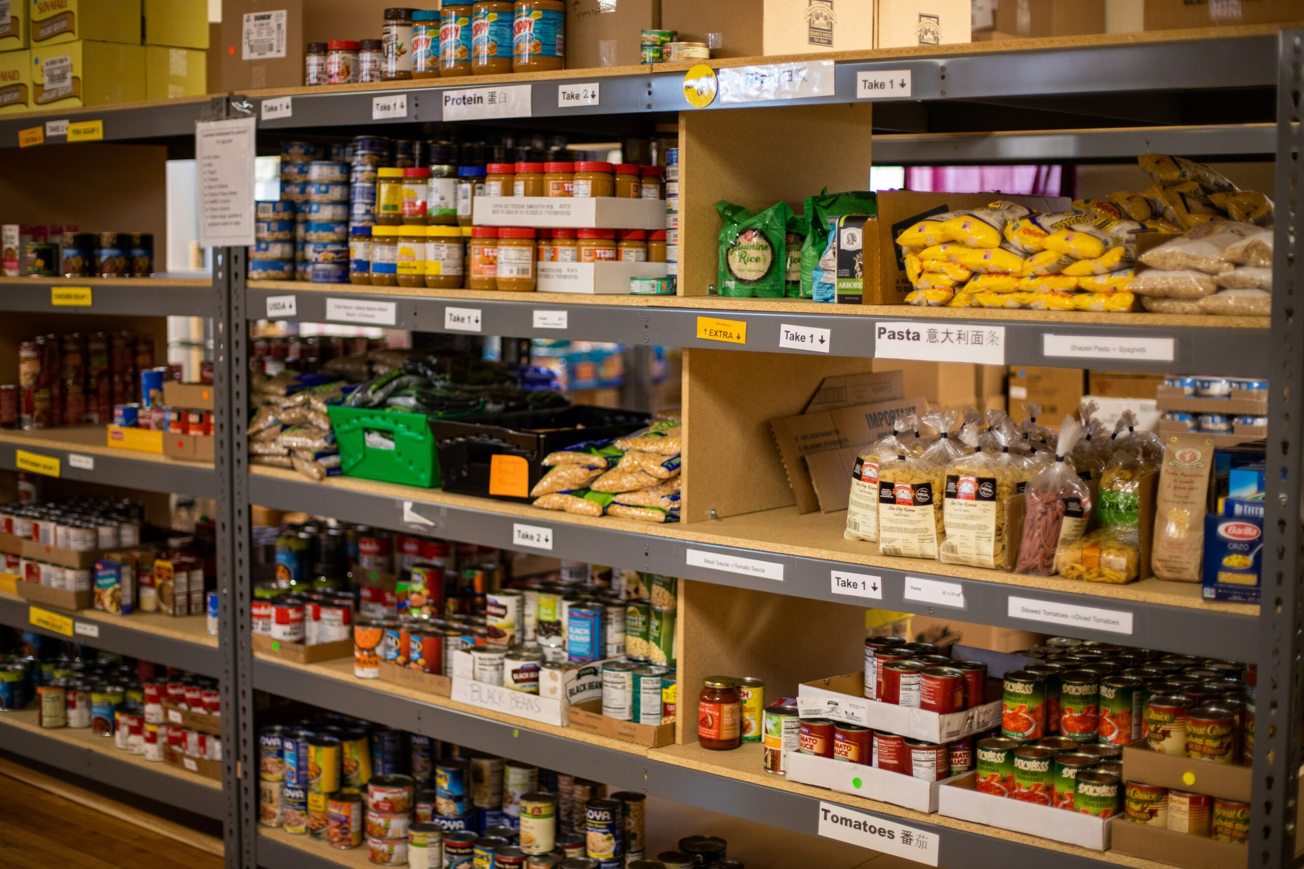 This is a photo of the inside of a food bank. No people can be seen. There are many shelves that have labels such as “pasta” “tomatoes” and “protein” and they also read “take one.”