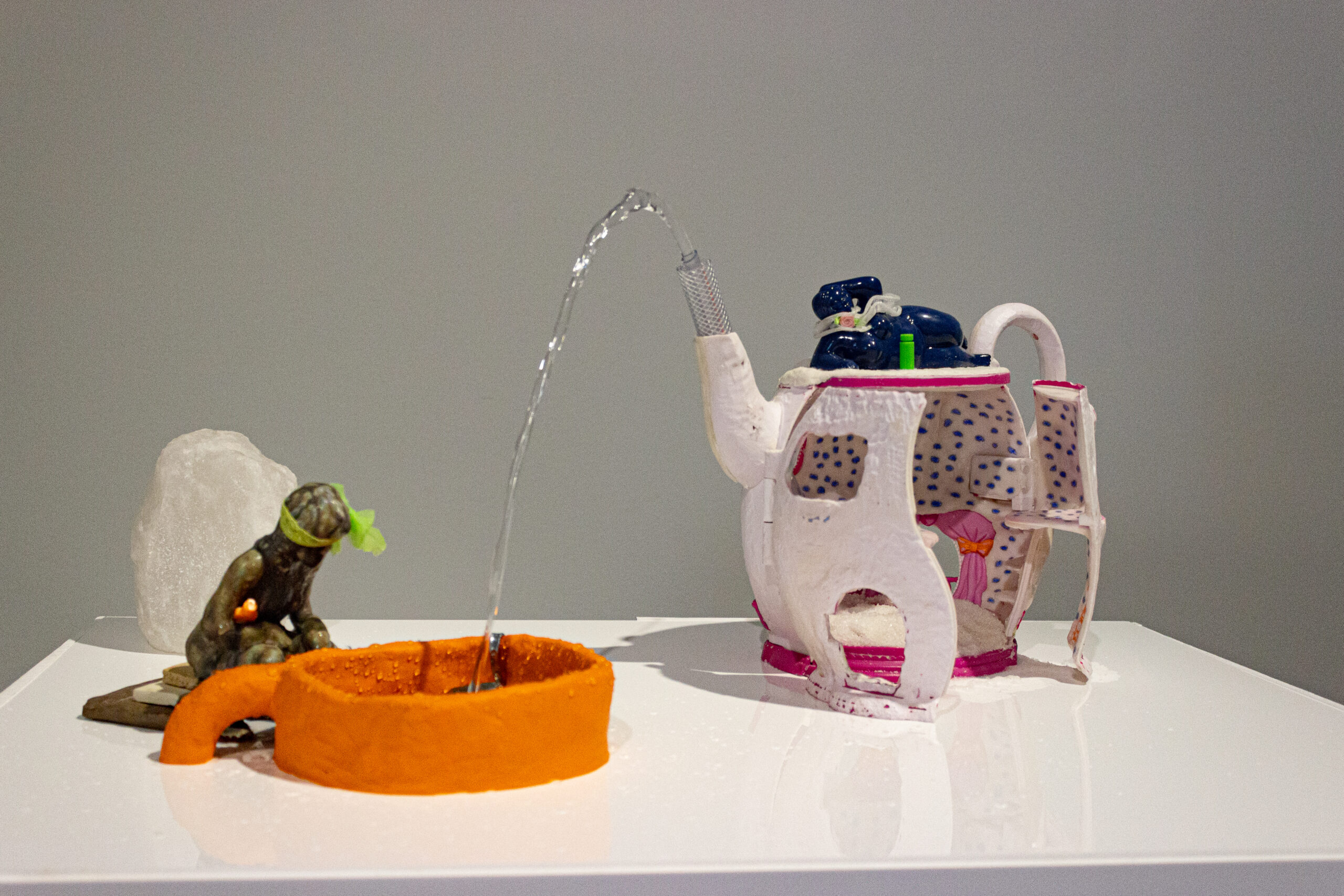 A sculpture of a white teapot with the front half open with windows like a dollhouse with the navy polka dots on the interior wallpaper. Water is coming out of the spout like a fountain, into an orange clay dish. Leaned over above the dish, a copper sculpture is kneeling over it.
