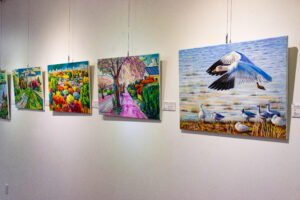 A row of four vibrant oil paintings hanging on the wall featuring surrealist depictions of trees, landscapes, and at the front, a white bird taking flight over a body of water.
