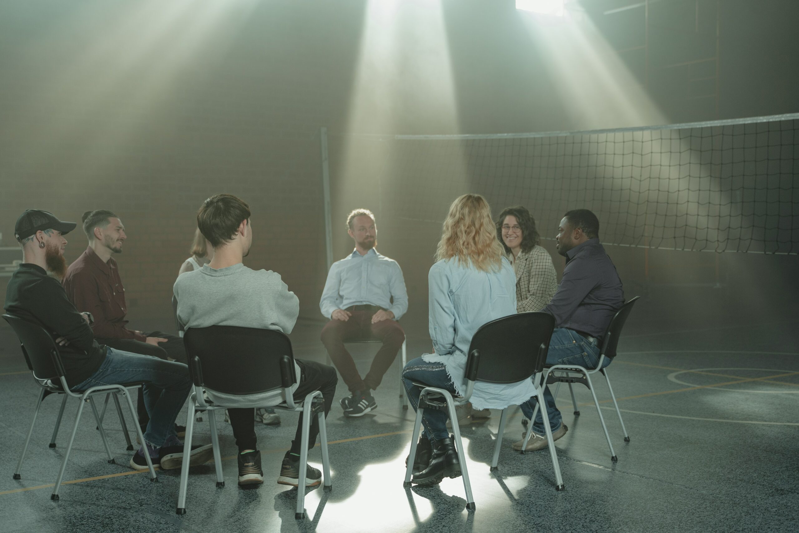 This is a photo of a group counselling session. Many people of diverse backgrounds are sitting in a circle of chairs in a large room.