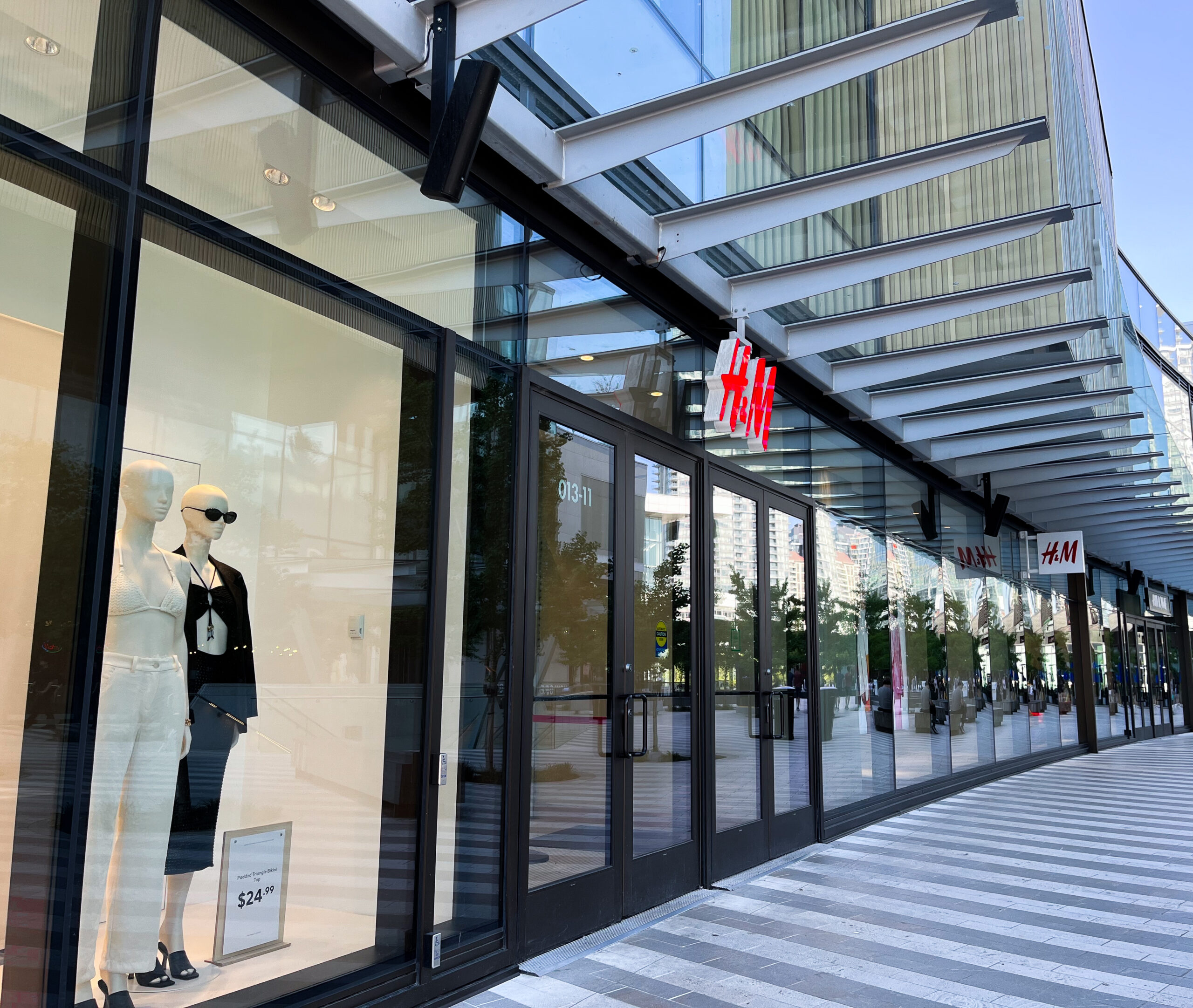 A modern-looking storefront exterior of H&M on a bright day. The display window features mannequins dressed in black and white monochrome outfits.