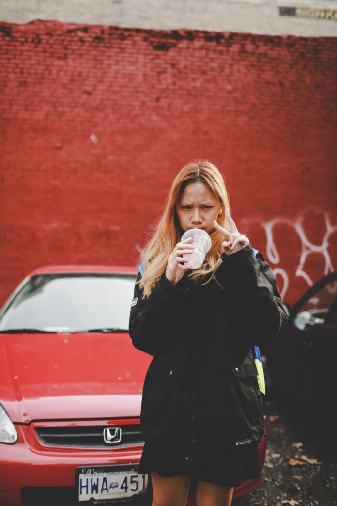 Woman holding a cup and making a peace sign with her hand beside a red car