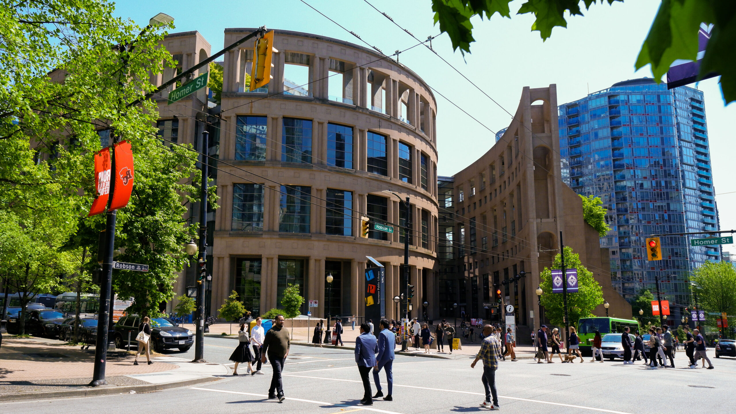 Photo of the exterior of Vancouver Public Library on Homer street on a sunny day with many pedestrians outside. The building’s architecture is a brown “rectangular box surrounded by a free-standing, elliptical, colonnaded wall.”